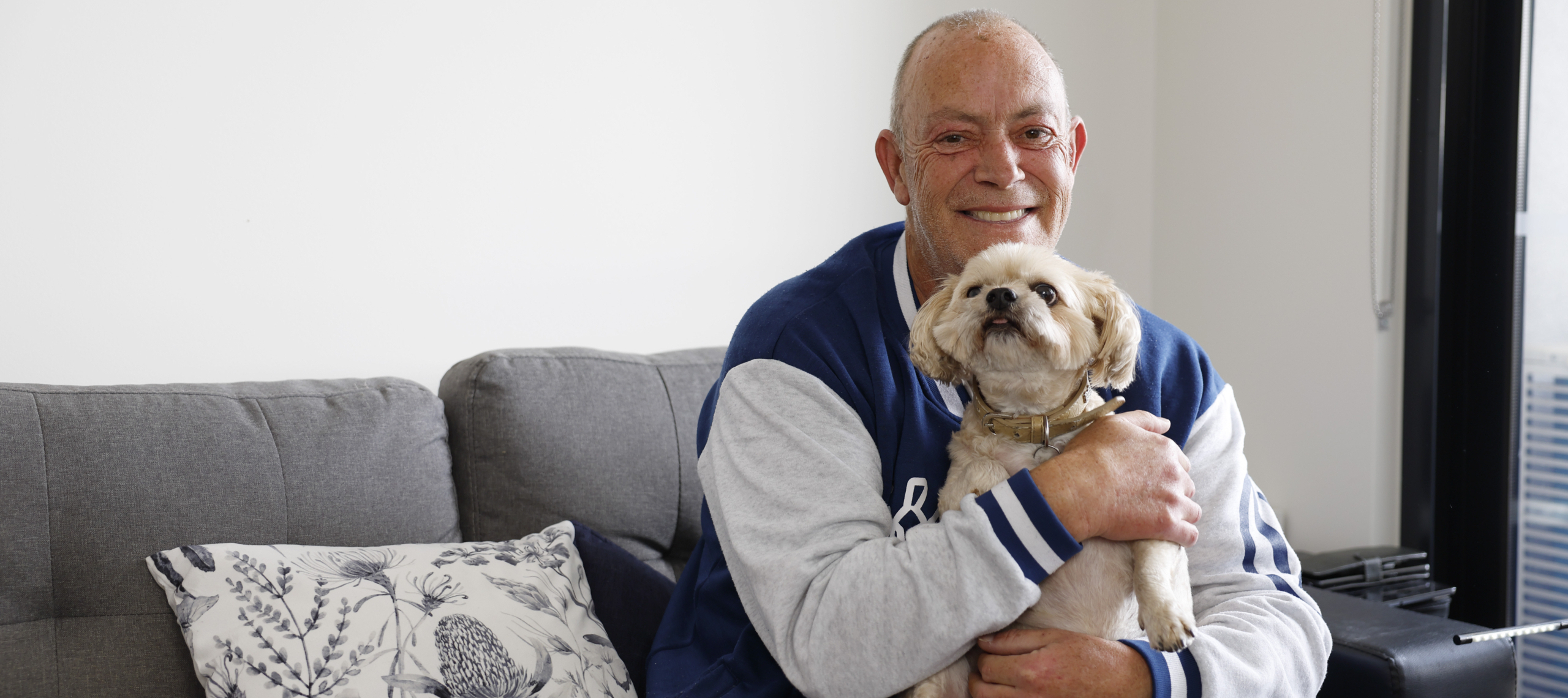 A smiling man in a blue and gray jacket sits on a gray couch holding a small, fluffy dog. There is a patterned pillow beside him and a large window in the background.