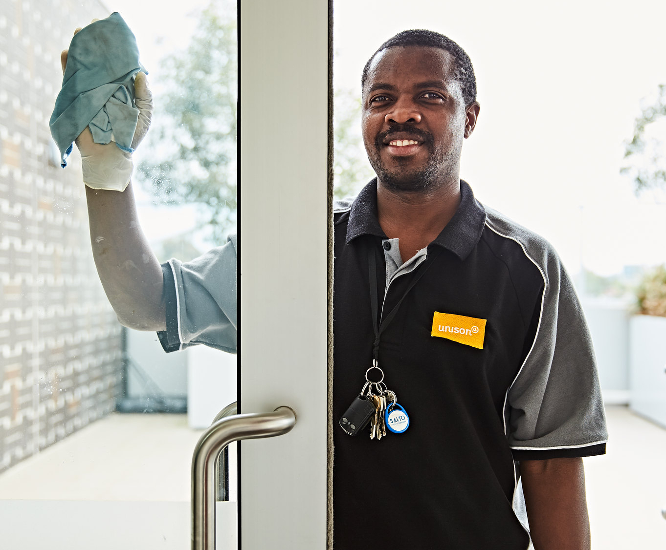 A man wearing a work uniform and gloves cleans a glass door with a blue cloth. He has keys and badges clipped to his shirt and is smiling at the camera.