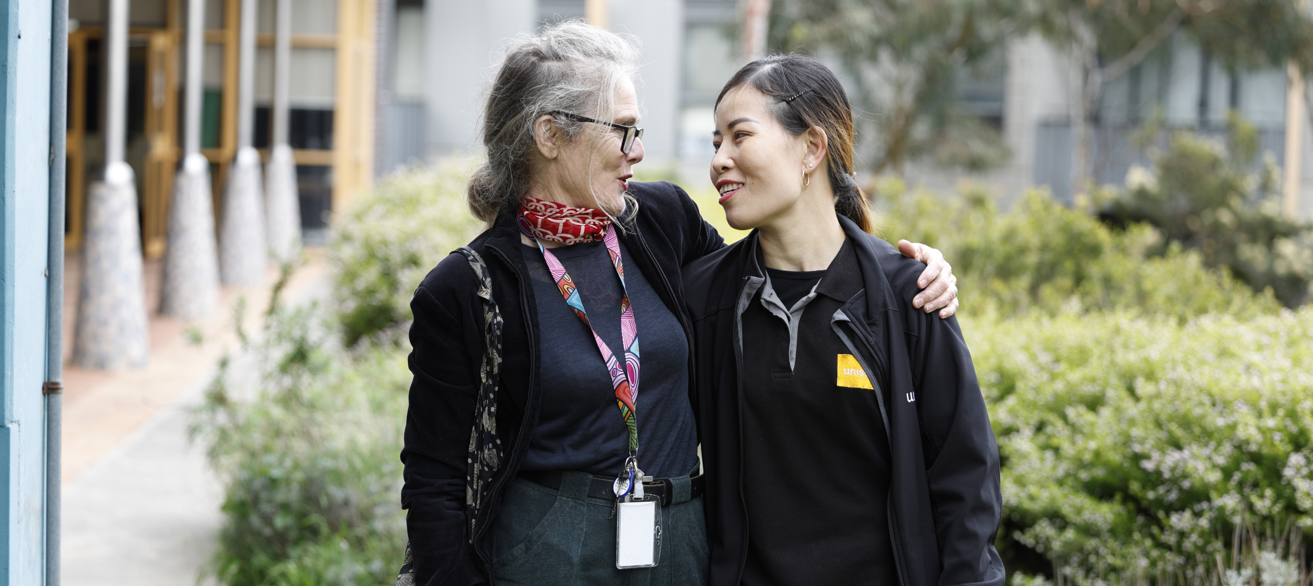 Two women stand outdoors, smiling at each other. One has gray hair and glasses, wearing a scarf and lanyard; the other has dark hair in a ponytail and wears a dark jacket. They have their arms around each other.