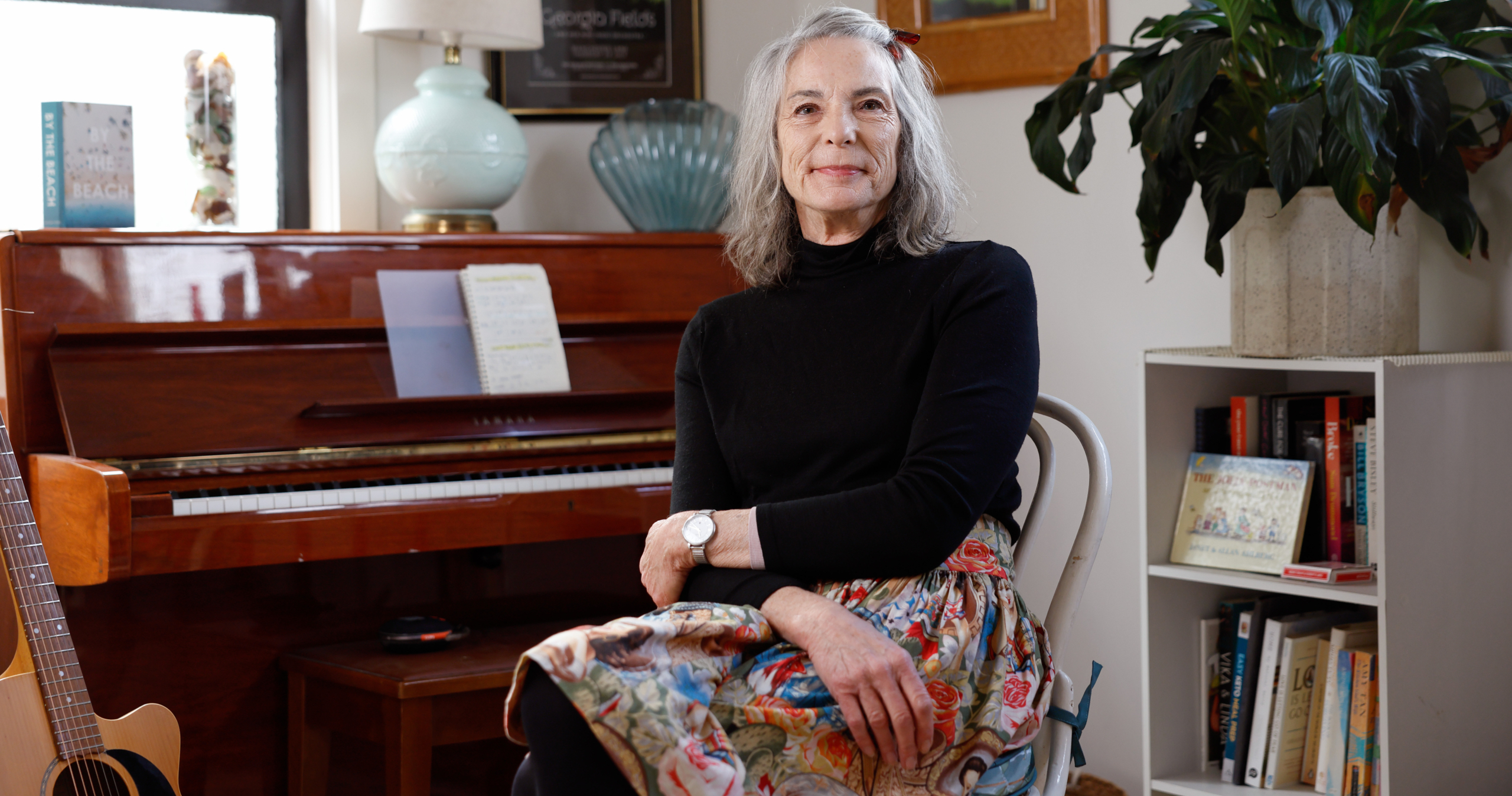 An older woman with gray hair, wearing a black top and floral skirt, sits on a chair in a cozy room with a piano, lamp, plants, books, and framed photos in the background. She is smiling gently at the camera.