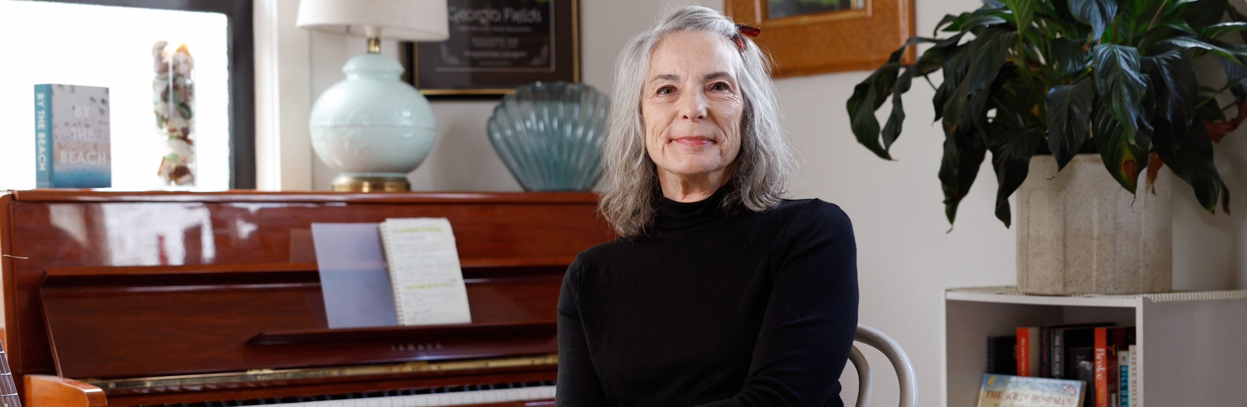 An older woman with gray hair, wearing a black top and floral skirt, sits on a chair in a cozy room with a piano, lamp, plants, books, and framed photos in the background. She is smiling gently at the camera.
