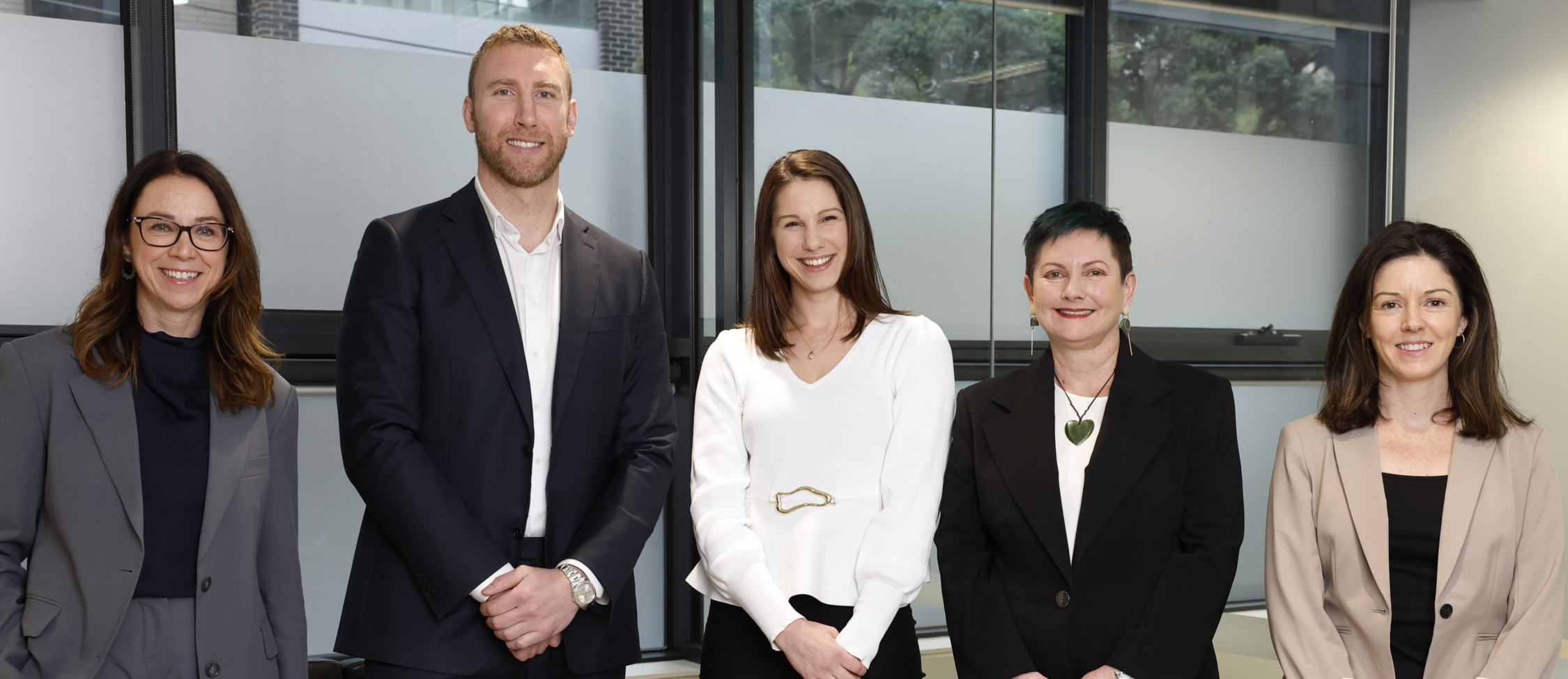 Five people in business attire stand side by side, smiling, in a modern office with large windows in the background.