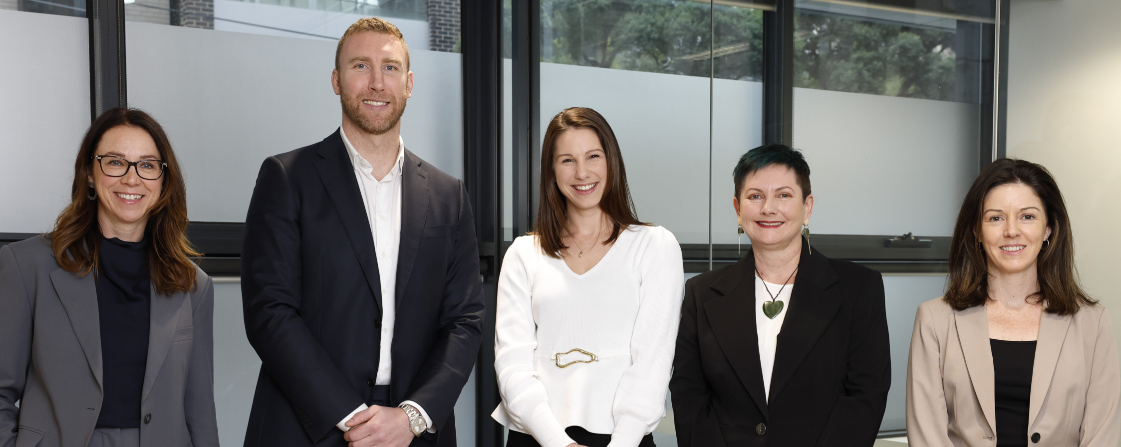 Five people in business attire stand side by side, smiling, in a modern office with large windows in the background.