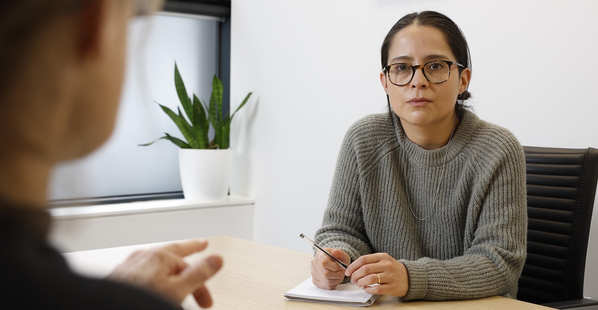 A person with glasses and a grey sweater sits at a desk, holding a pen and notepad, listening attentively to someone speaking off-camera. A green potted plant is on the windowsill in the background.