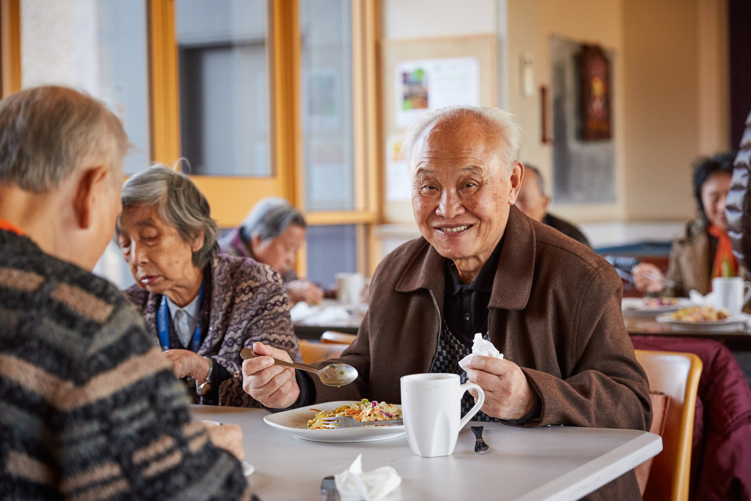 An elderly man smiles while eating a meal at a table with other seniors in a communal dining setting. He holds a fork and napkin, and other people are also enjoying their food in the background.