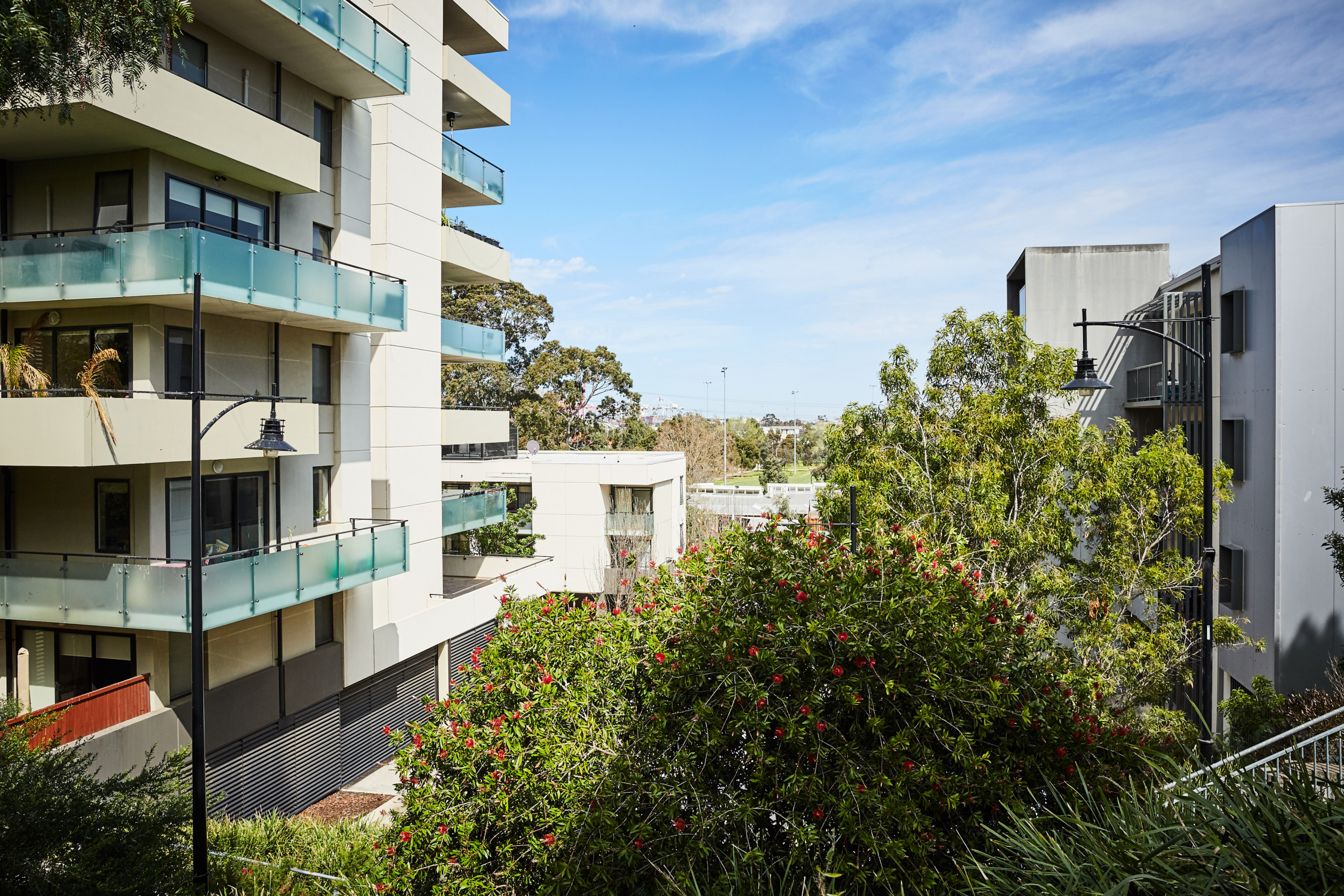 Modern apartment buildings with glass balconies frame a lush green garden with red flowers in the foreground, under a partly cloudy blue sky. Trees and more buildings can be seen in the background.