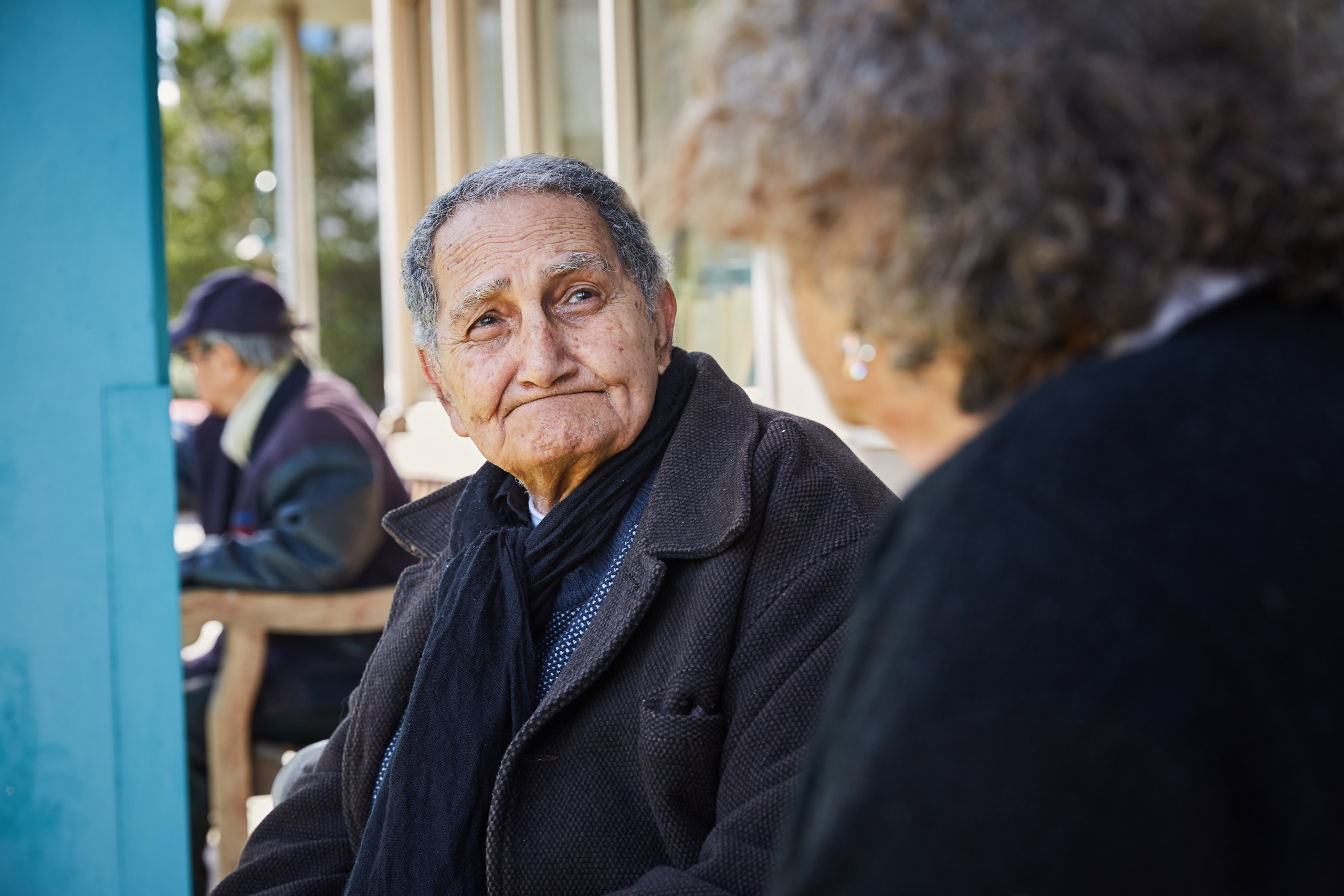 An elderly man wearing a dark coat and scarf sits on a bench outdoors, looking thoughtfully at another person in the foreground. Another person is seated in the background.