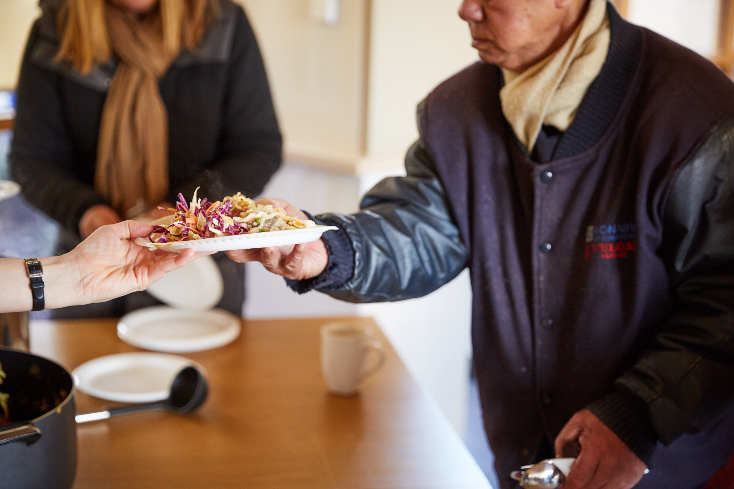 A person hands a plate of food to another person wearing a scarf and jacket, suggesting a meal being served in a community or charitable setting. A pot and mug are visible on the table.