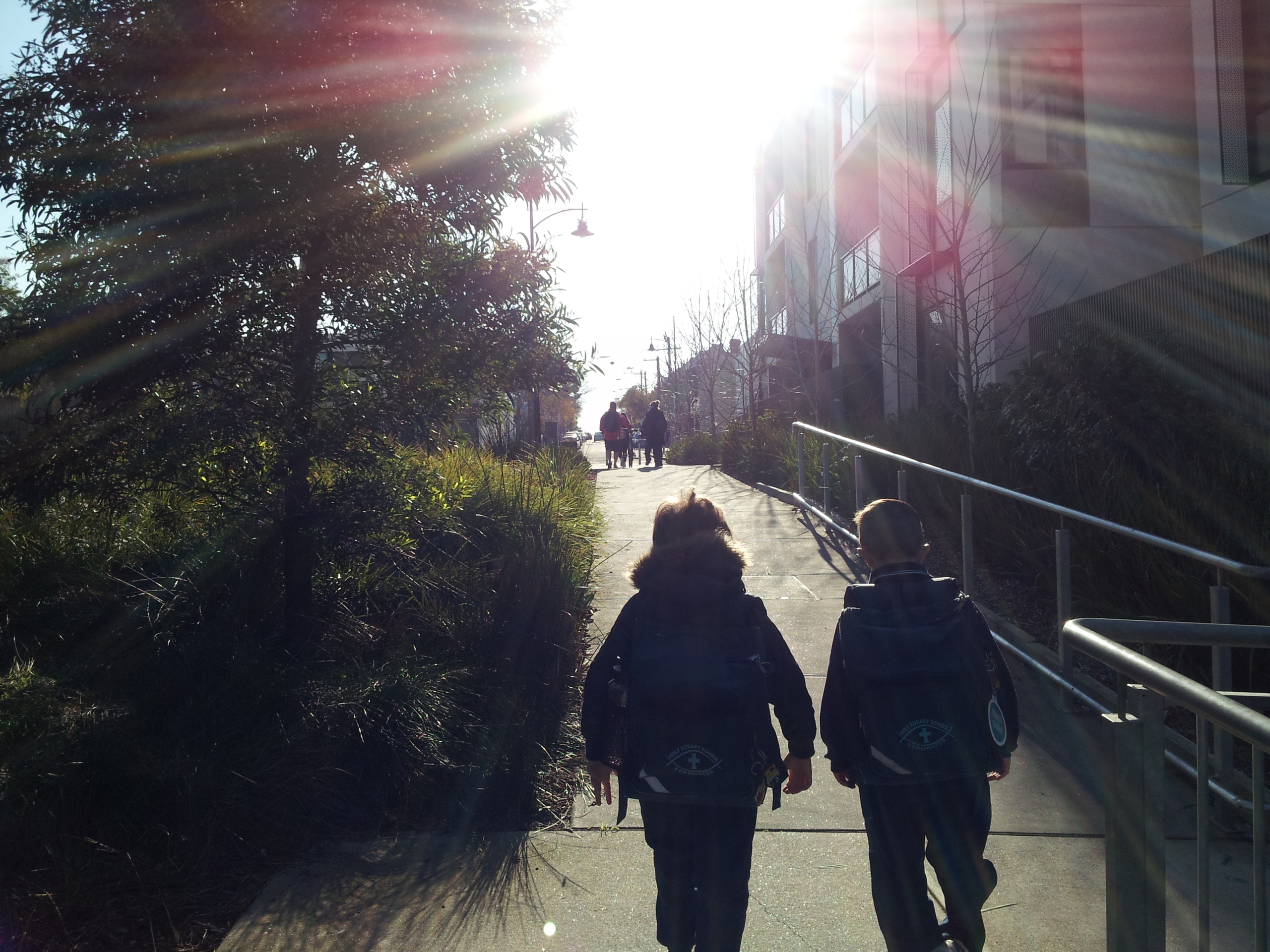 Two children walk up a sunlit path, holding hands, with the sun shining brightly overhead. Shadows and greenery line the walkway, and several people are visible in the distance.