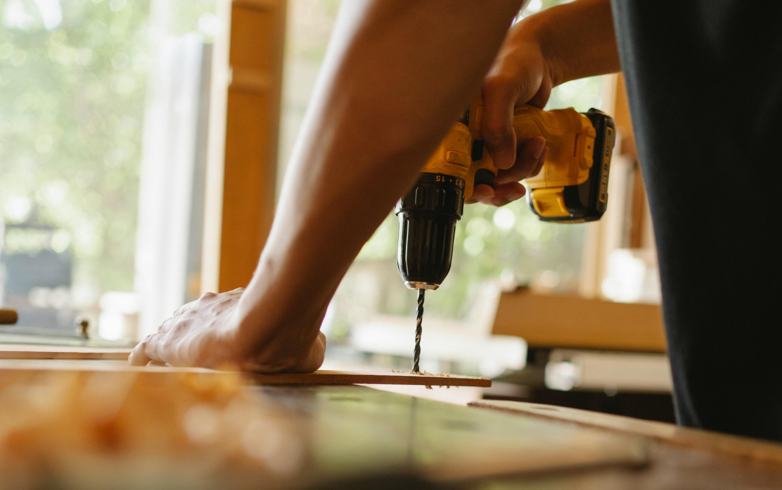 A person uses a yellow power drill to make a hole in a wooden board, holding the wood steady with one hand in a well-lit workshop.