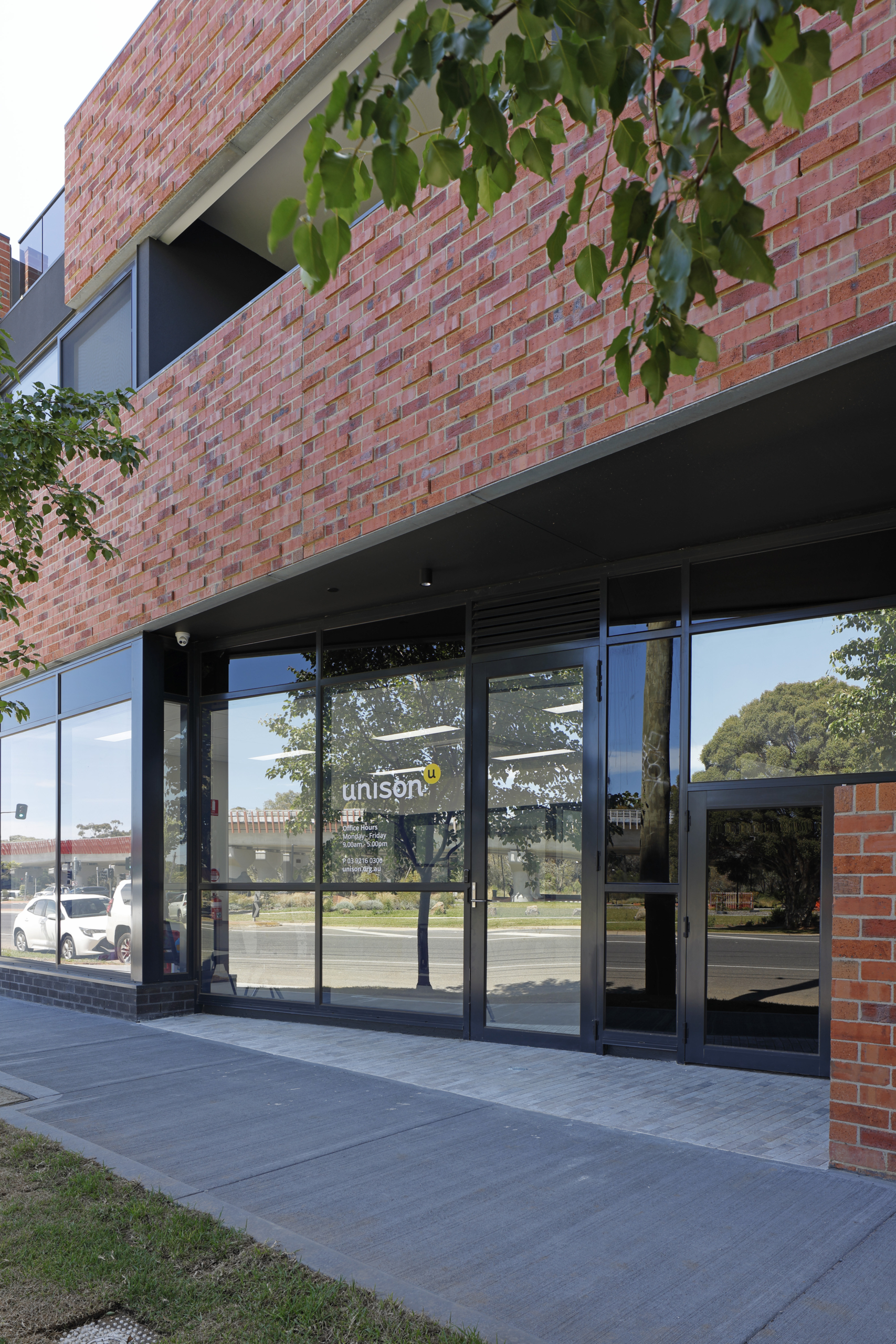 A modern building with red brick exterior and large glass windows at street level. Reflections of trees and cars are visible in the glass, and a sign on the door reads unison. A sidewalk and green leaves are in the foreground.