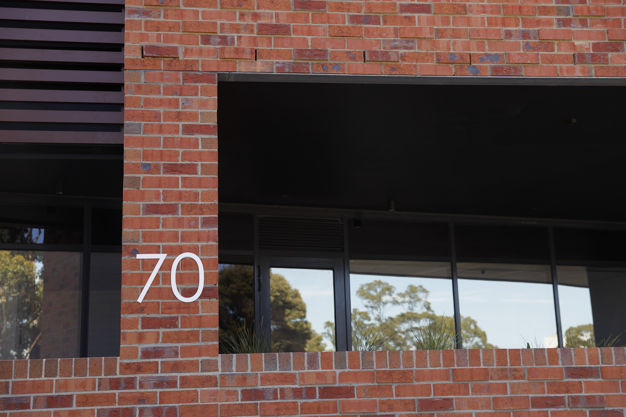 A modern red brick building with large black-framed windows and the number 70 displayed in white on the exterior wall. Trees are reflected in the window glass.