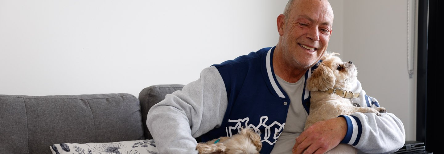 A smiling man sits on a gray couch, hugging a small tan dog while another dog rests beside him. He wears a blue and gray jacket, and the scene appears warm and relaxed in a home setting.