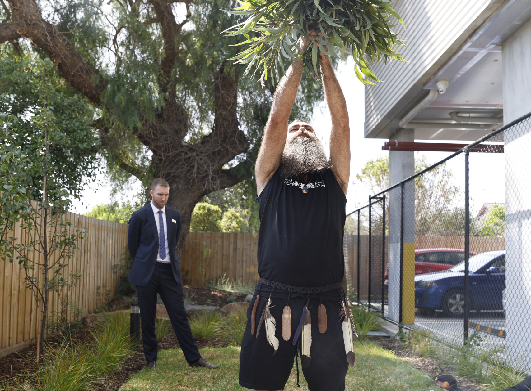 A man with a long beard, wearing a black shirt and shorts with a traditional belt of pouches, lifts a bundle of greenery overhead during an outdoor ceremony. Another man in a suit stands in the background, watching.