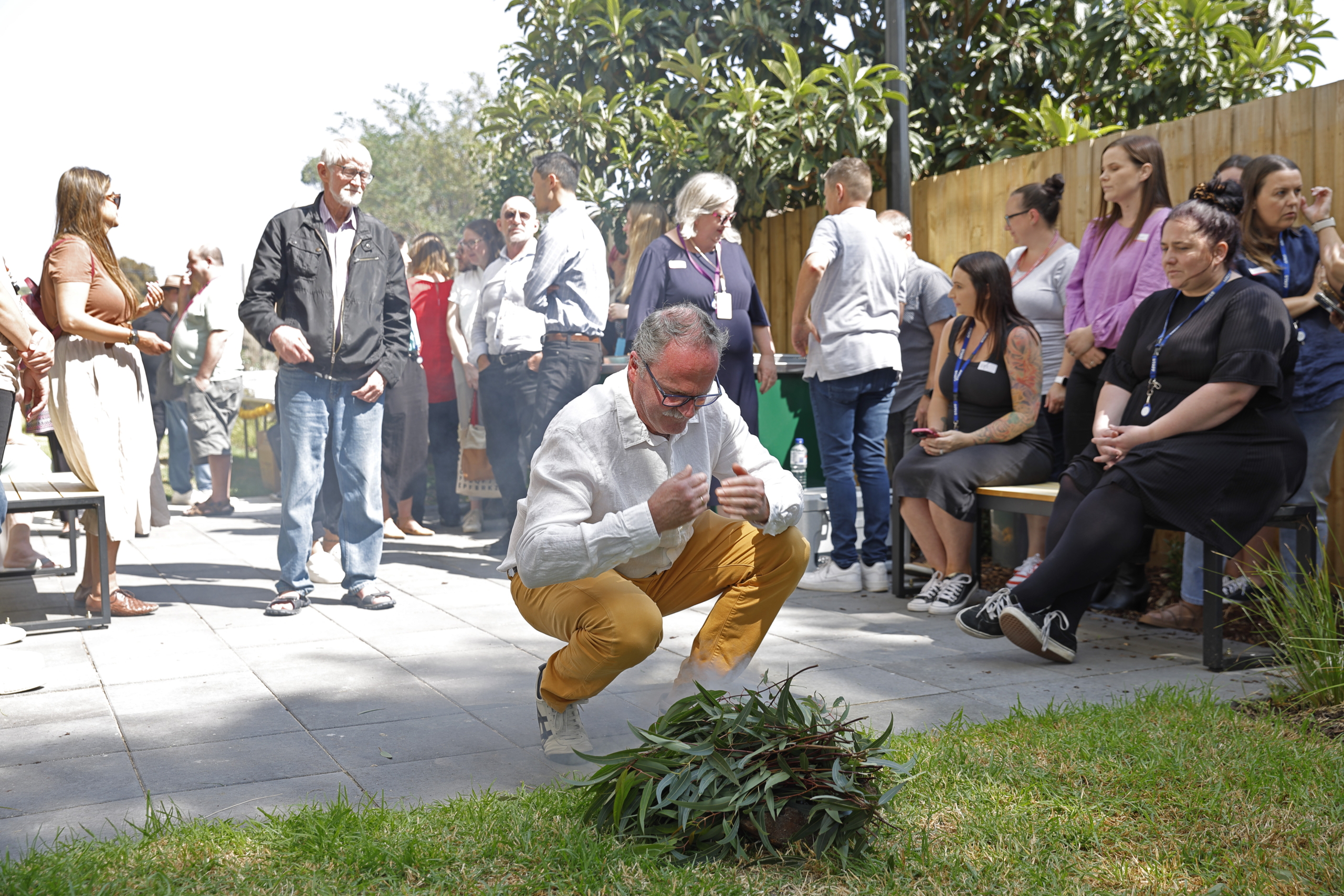 A man crouches near a small pile of greenery, possibly for a traditional smoking ceremony, while a diverse group of people watch and gather outdoors in a courtyard.